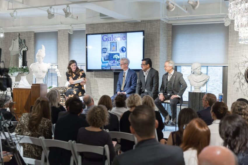 Panel discussion at an art event with four speakers on stage and an audience, statues and a screen in the background.
