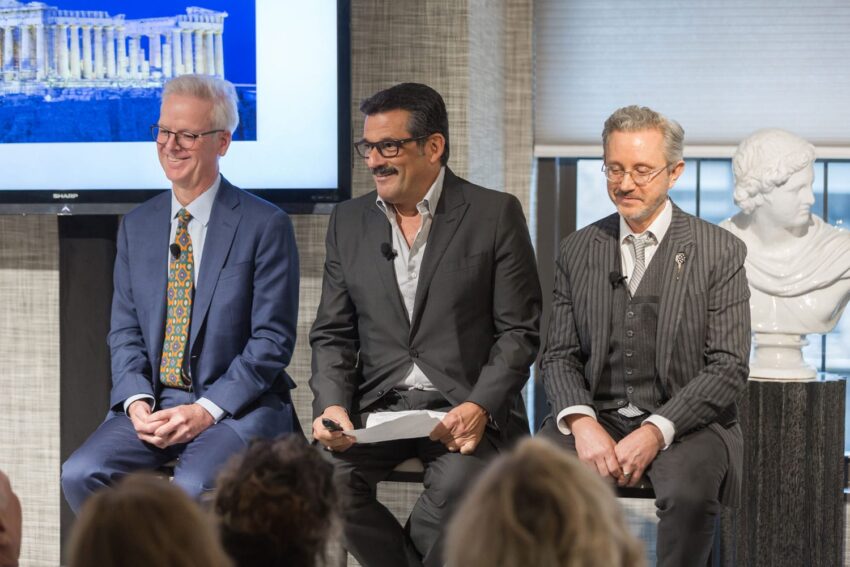 Three men seated on stage in suits during a speaking event with a screen and a statue in the background.