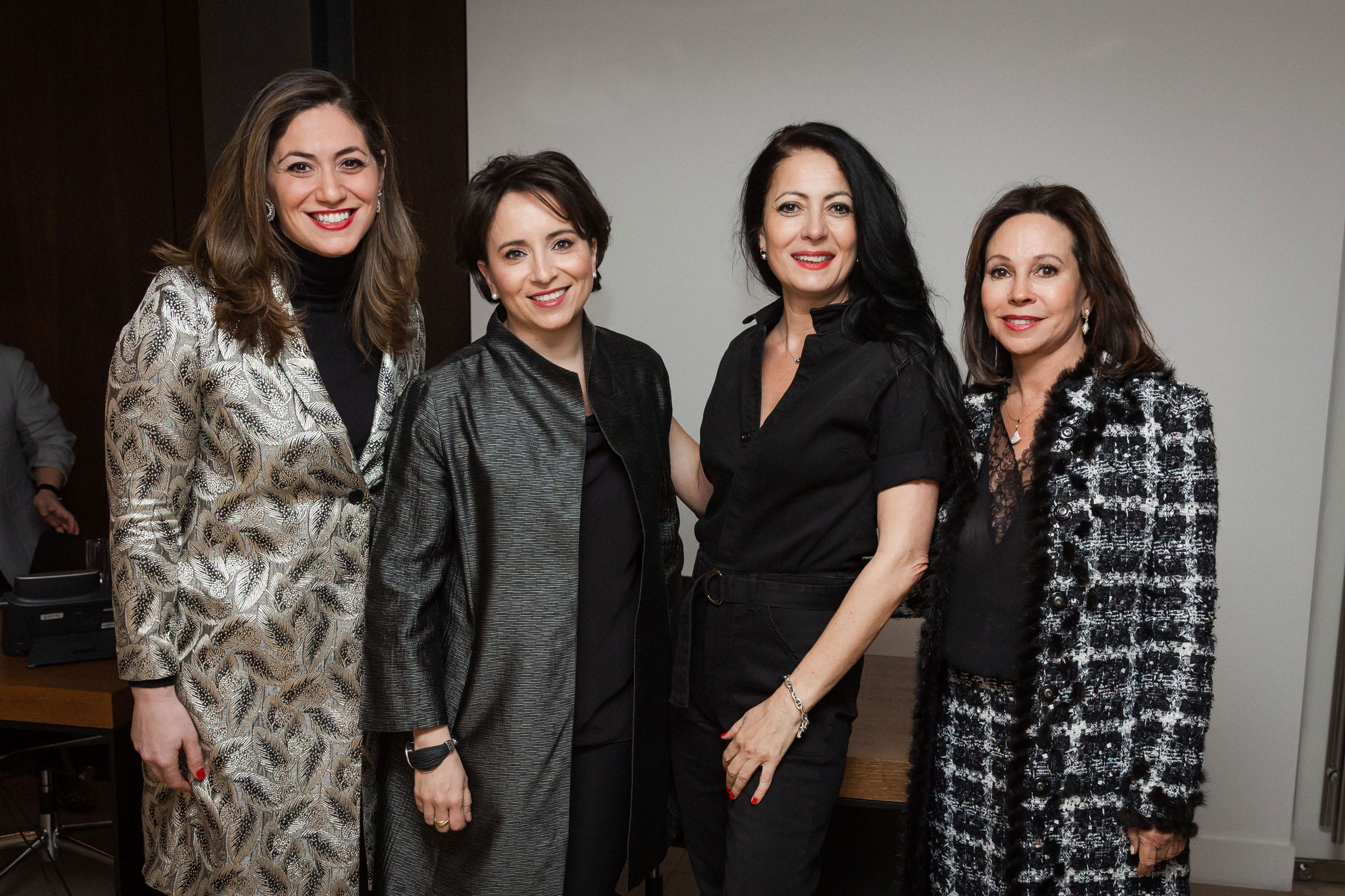 Four women posing together, smiling, indoors, wearing stylish outfits.