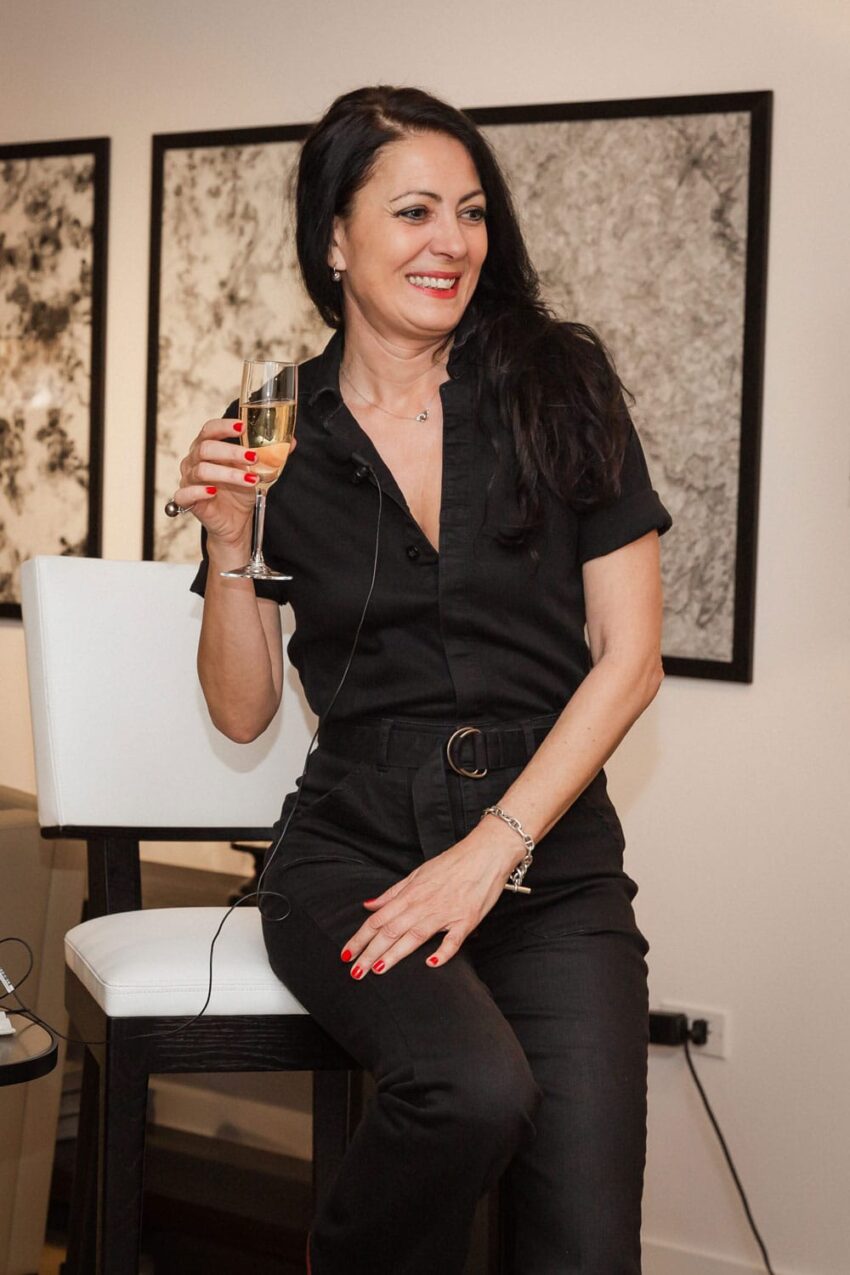 Woman in a black outfit sitting on a chair holding a glass, smiling at an indoor event.