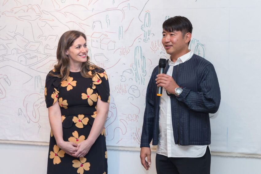 Two people smiling, standing in front of a whiteboard with a drawing. One is speaking into a microphone.