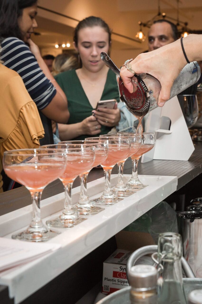 Bartender pouring cocktails into glassware on a bar counter with people gathered in the background.