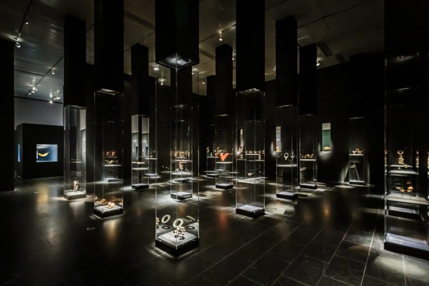 Dimly lit museum gallery displaying ancient gold artifacts in glass cases.