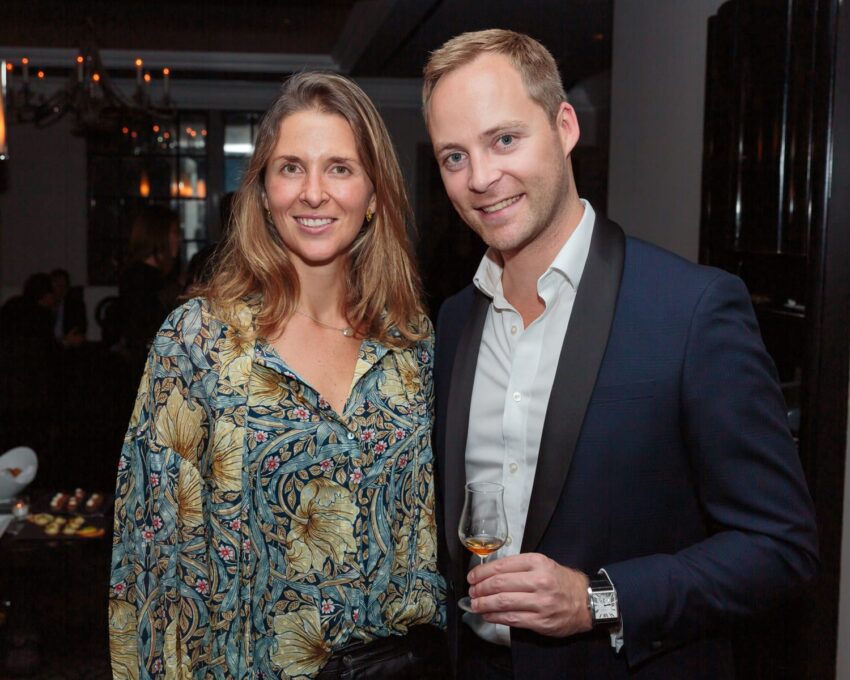 Smiling man and woman at an event, man holding a glass, indoor setting with warm lighting.