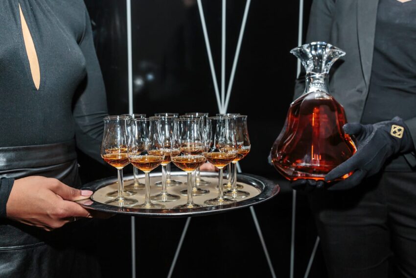 Two people holding a tray of filled wine glasses and a decorative decanter against a dark background.