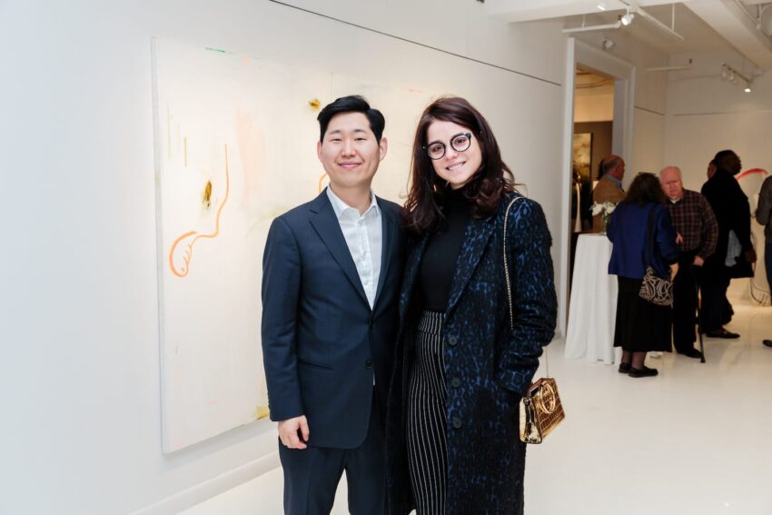 Two people smiling in an art gallery with abstract painting and guests in the background.