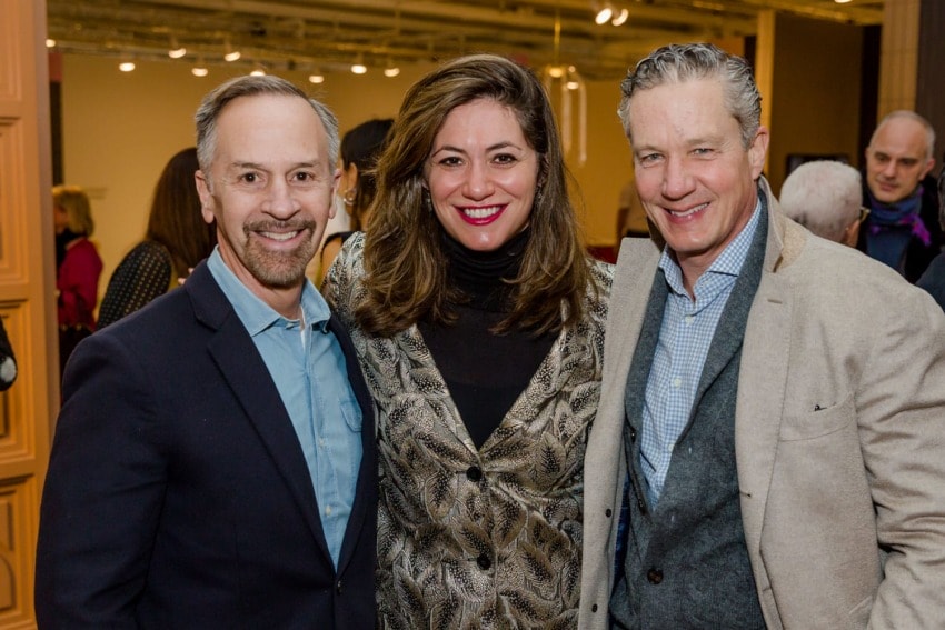 Three people smiling at a gathering, standing inside a well-lit room with a patterned wall in the background.