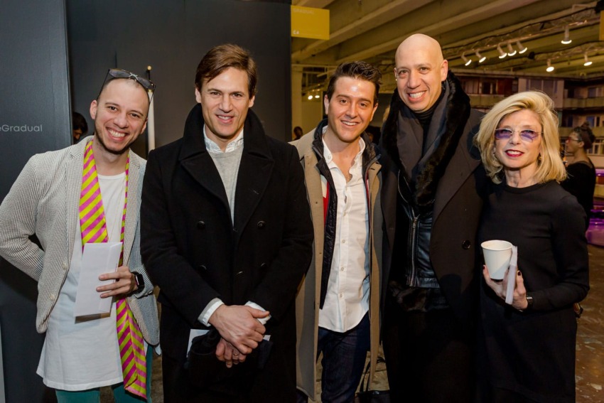 Group of five people smiling and posing together indoors, wearing stylish outfits, one holding a coffee cup.