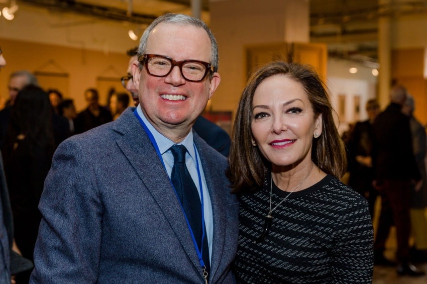 Man in glasses and blazer stands smiling with a woman in a black dress at a social event indoors.