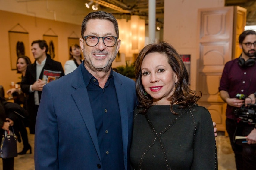 Smiling man in suit and glasses with woman in black dress at indoor event with people in the background.
