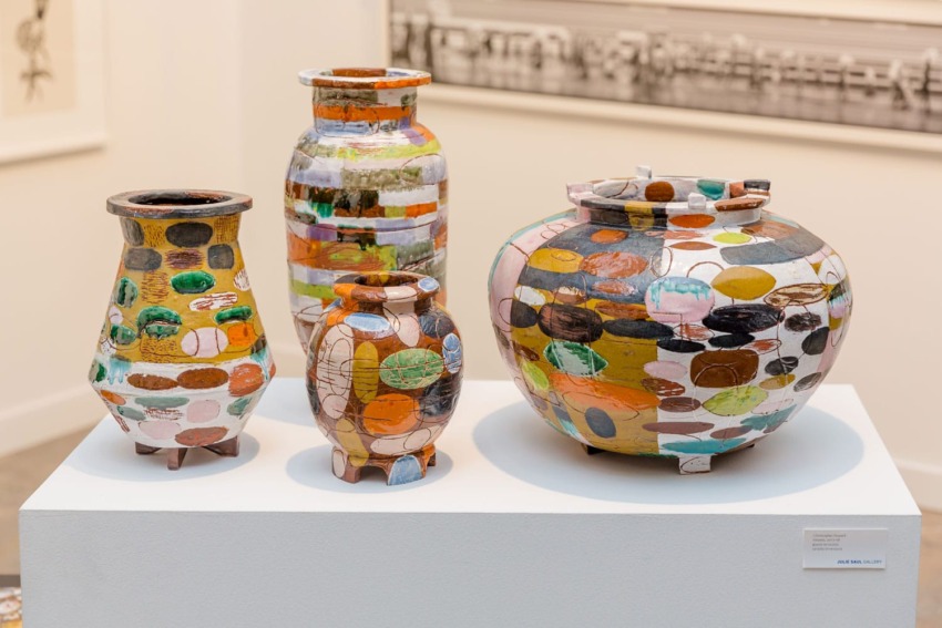 Colorful, painted ceramic pots of various sizes displayed on a white pedestal in an art gallery setting.
