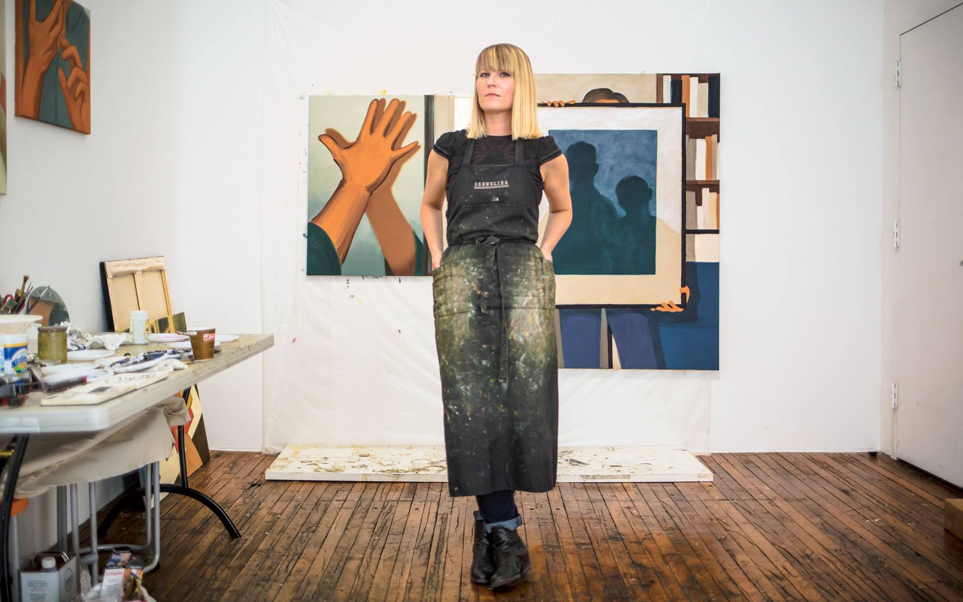 Artist standing in studio with paintings and art supplies visible, wearing an apron and looking contemplative.