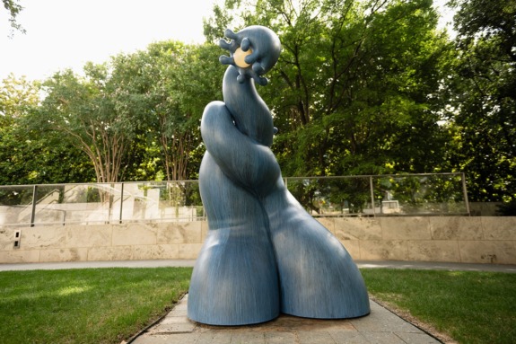 Blue abstract sculpture resembling draped figures, set outdoors against lush green trees and stone pathway in a park setting.