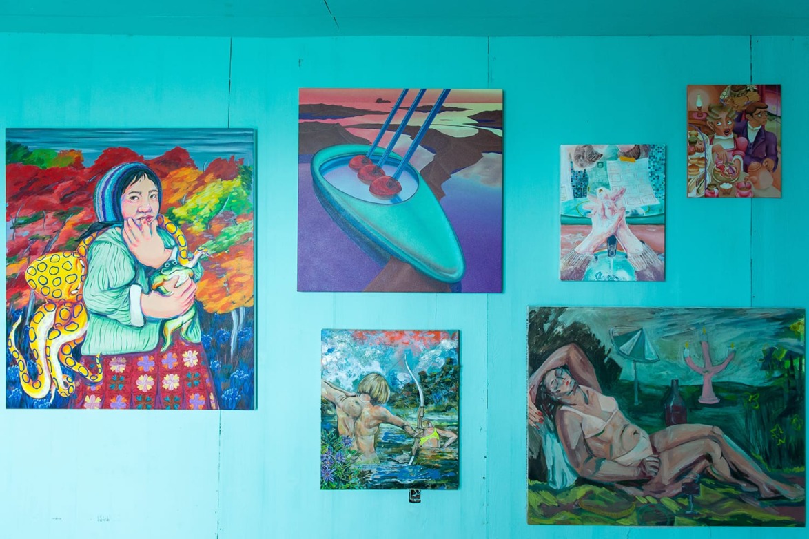 Colorful paintings displayed on a turquoise wall, featuring various subjects including people, nature, and abstract designs.
