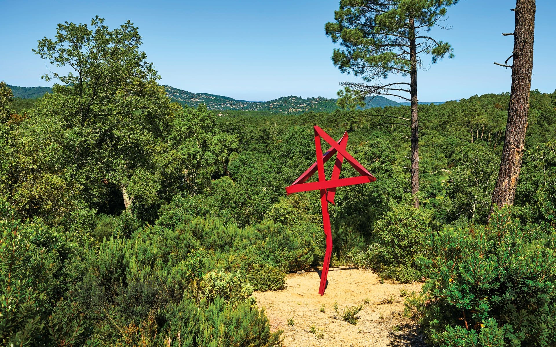 Red abstract sculpture in a lush forest setting under a clear blue sky, with distant hills visible in the background.