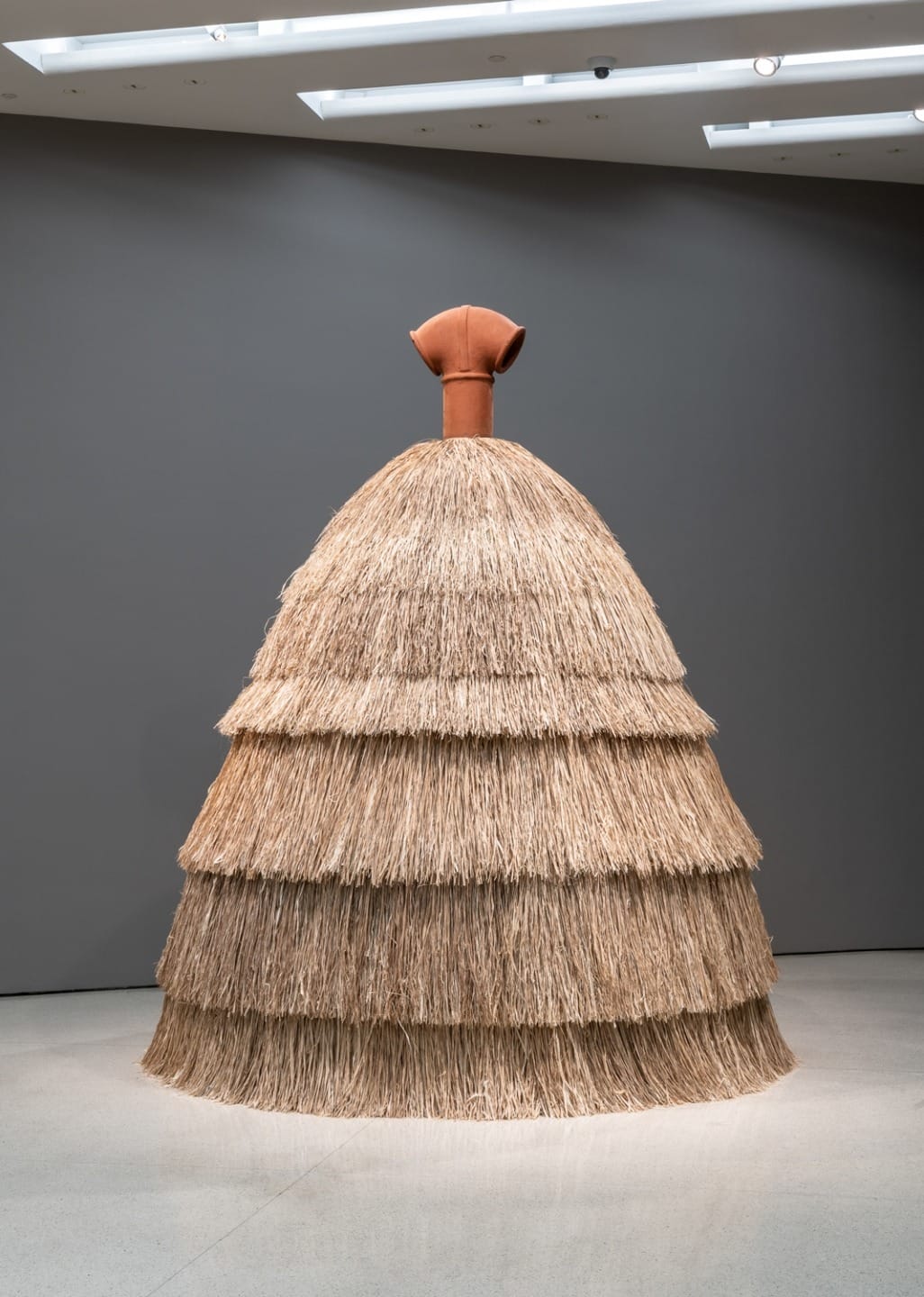 Art sculpture resembling a dress with layered straw skirt and a terracotta-colored top, displayed in a modern gallery space.
