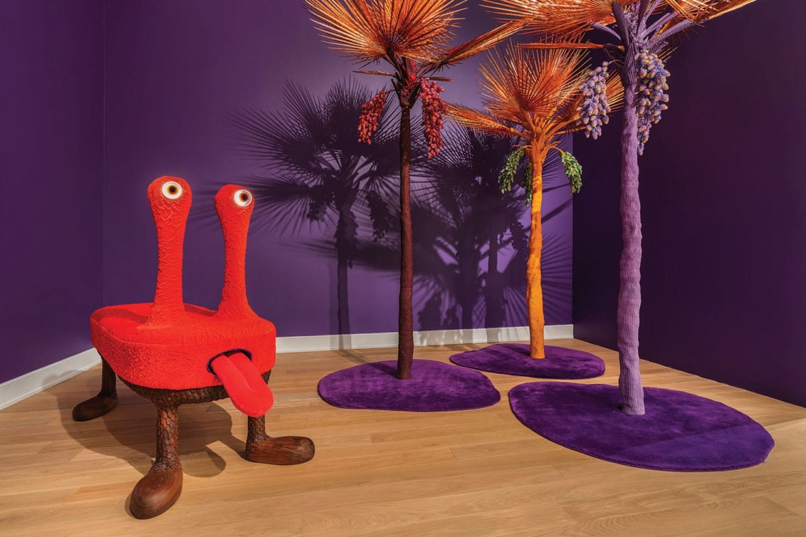 Quirky red creature chair with eyes next to vibrant purple palm trees on a wooden floor against a purple wall.