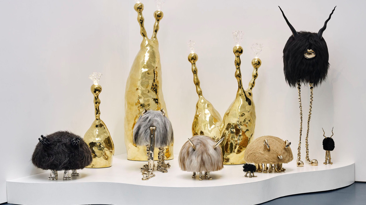 Sculptural art display with metallic and furry abstract figures on a white platform against a plain background.