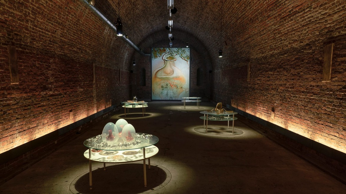 Art installation in an arched brick gallery with round glass tables displaying various sculptures under warm lighting.