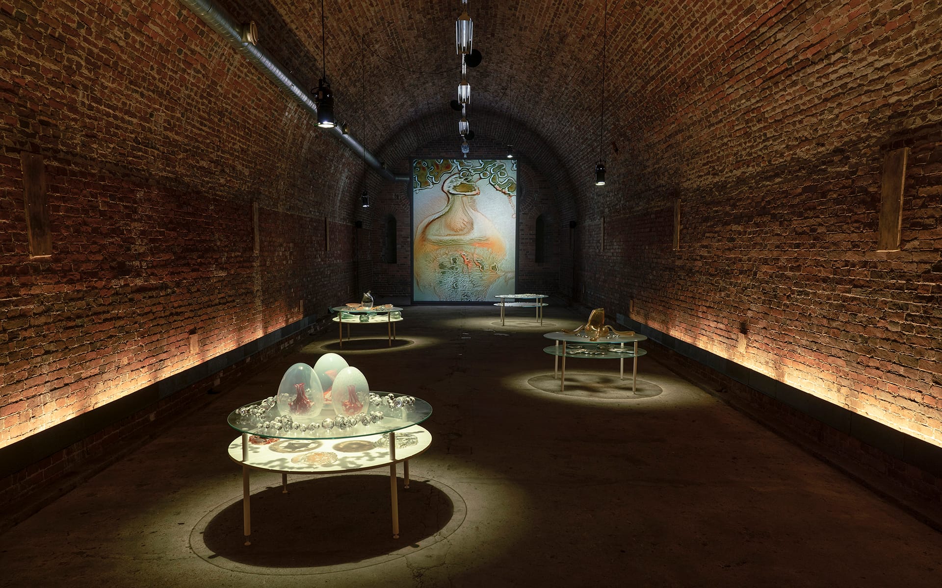Art installation in an arched brick gallery with round glass tables displaying various sculptures under warm lighting.