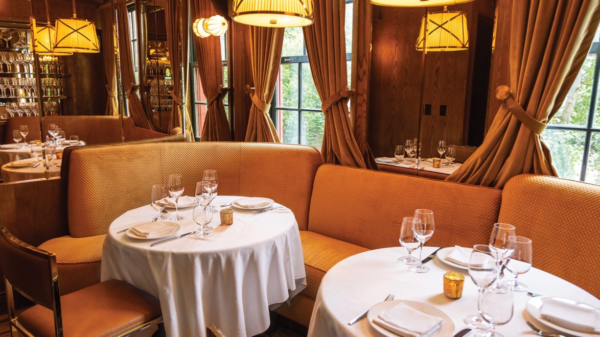 Cozy restaurant interior with elegant table settings, round tables covered in white tablecloths, and warm lighting.