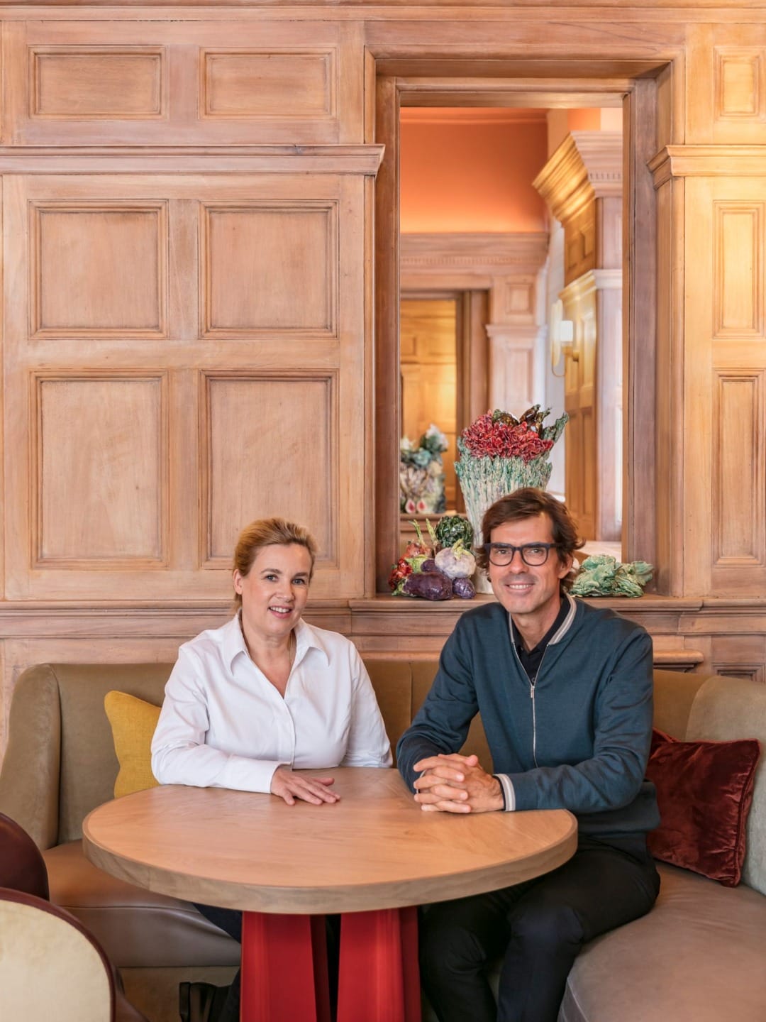 Two people sitting at a round table in a warmly decorated room with wooden paneled walls and floral arrangements in the background.