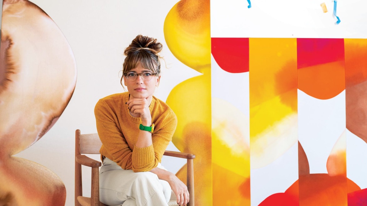 Person with glasses sitting on a chair, surrounded by colorful abstract art pieces in an art studio.
