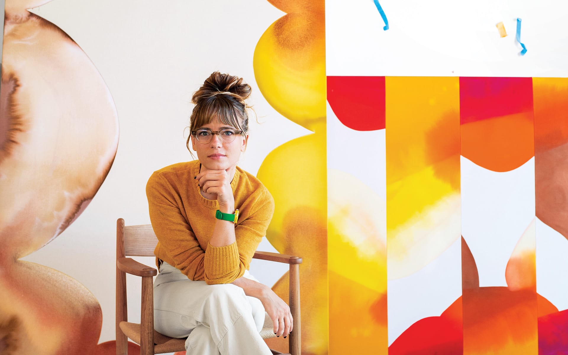 Person with glasses sitting on a chair, surrounded by colorful abstract art pieces in an art studio.