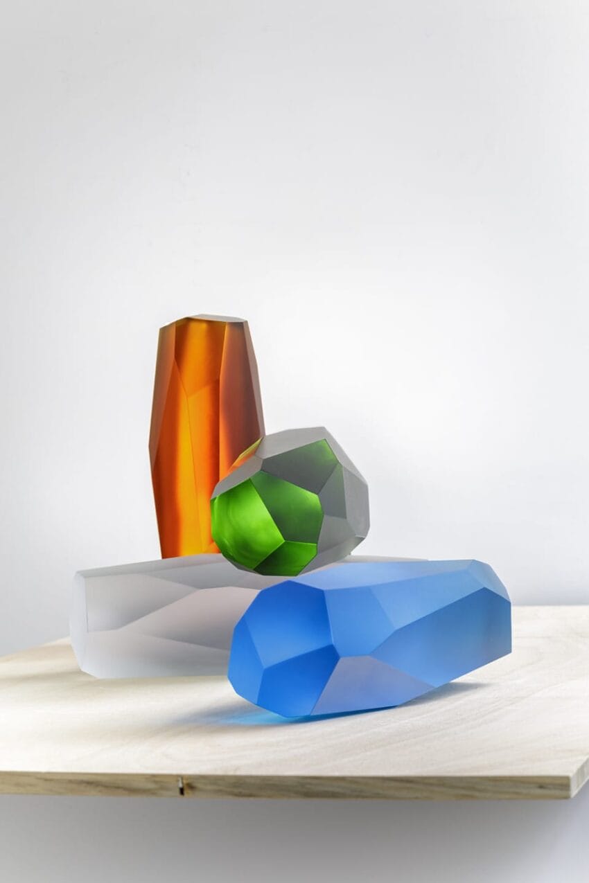 Geometric glass sculptures in orange, green, blue, and clear on a wooden surface against a white background.