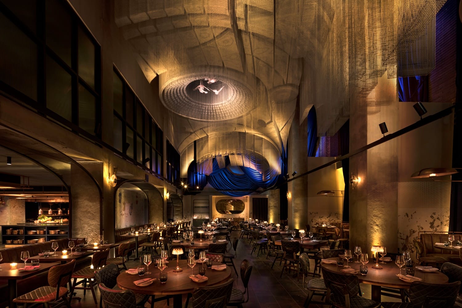 Dimly lit elegant restaurant with high ceilings, warm lighting, and tables set with glasses, silverware, and plates.