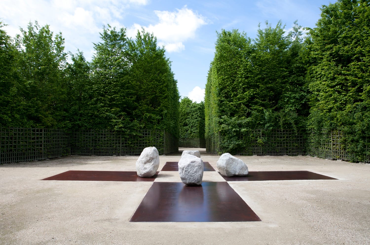 Garden pathway with three large stone sculptures and geometric designs surrounded by lush green hedges under a blue sky.