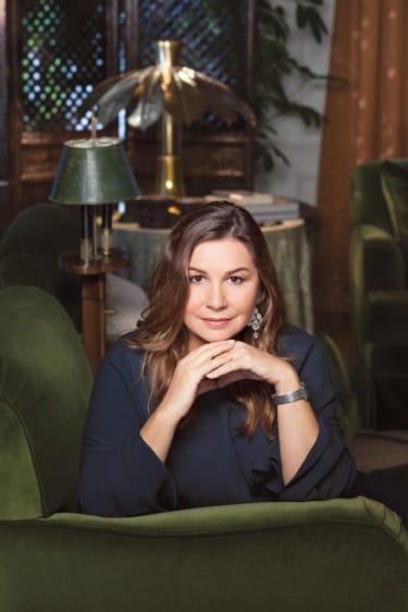 Woman sitting on a green couch in a cozy living room with warm lighting and elegant decor.