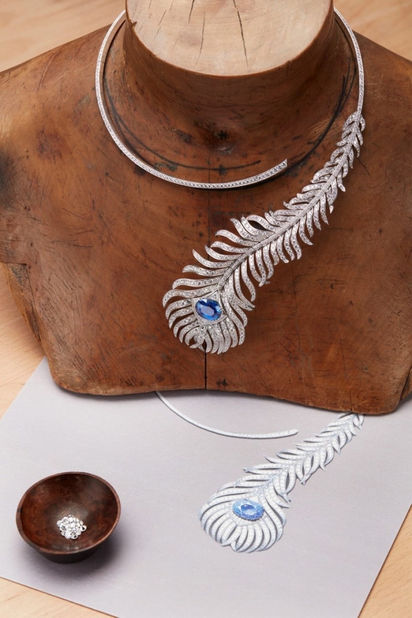 Diamond and sapphire feather necklace displayed on wooden stand with matching jewelry design sketch and loose gems nearby.