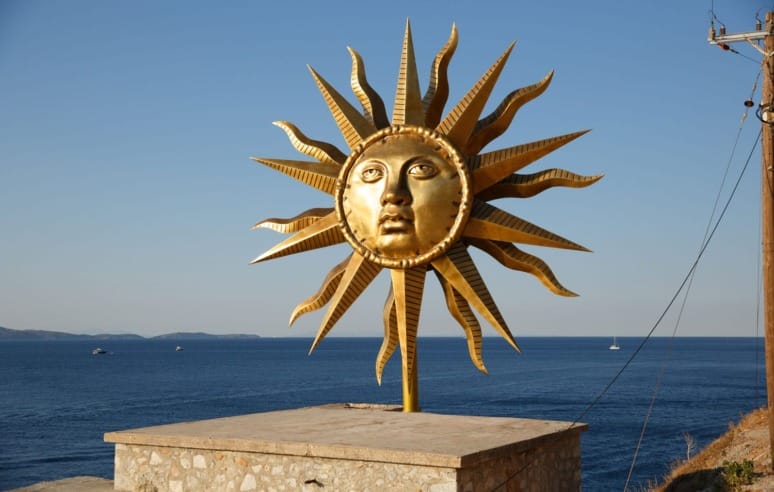Golden sun sculpture with a face, featuring radiating sunbeams, set against a clear blue sky and sea backdrop.
