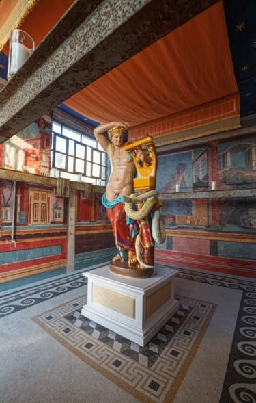 Colorful statue of a seated figure holding a lyre in a richly decorated room with vibrant murals and geometric floor patterns.