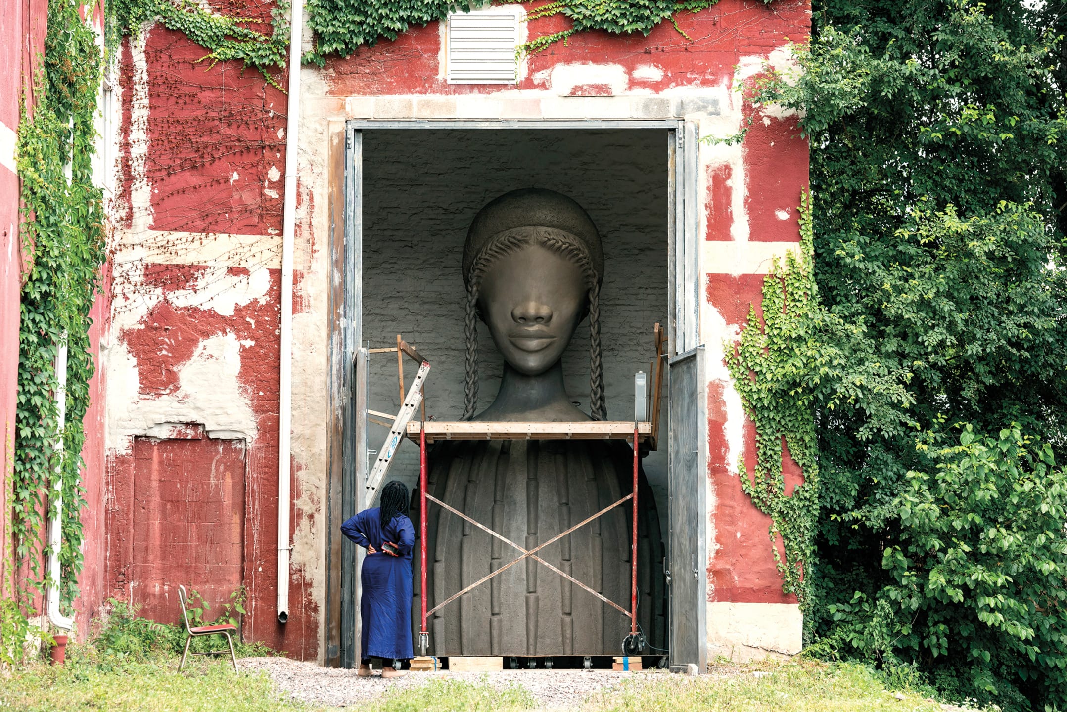 Person in blue robe observes a large sculpture of a woman's head inside a building with ivy-covered walls.