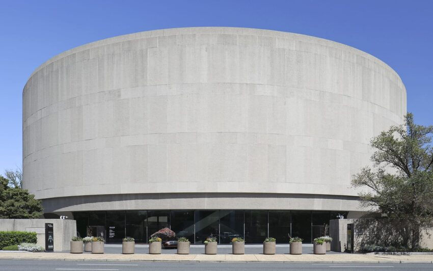 Exterior view of a large circular concrete building with glass entrance doors and planters, set against a clear blue sky.