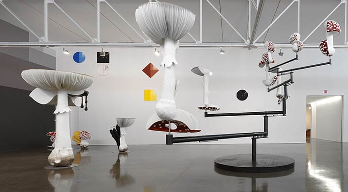 Modern art installation with large mushroom sculptures and geometric shapes in a white-walled gallery space.