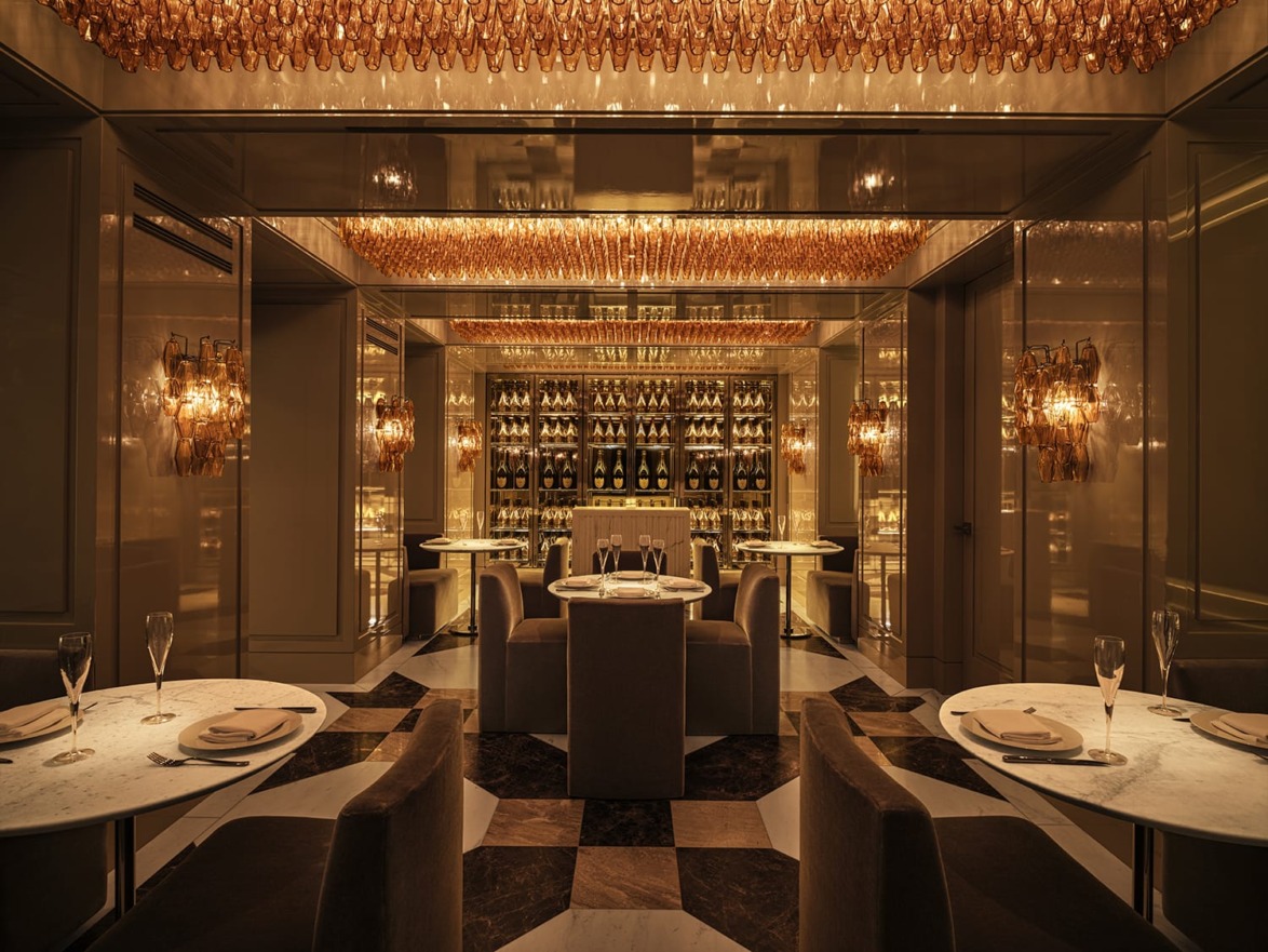 Elegant restaurant interior with marble tables, plush seating, and a wall display of champagne bottles, warmly lit ambiance.