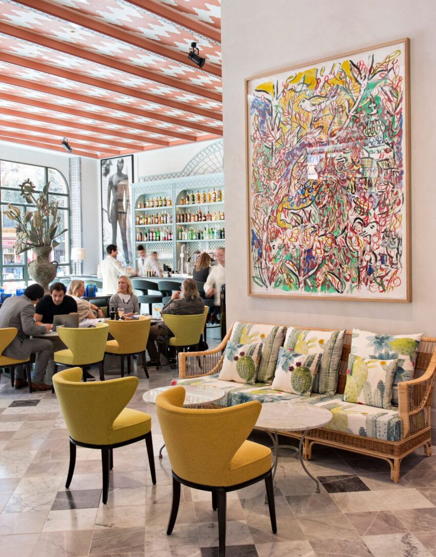 Modern cafe with colorful artwork, yellow chairs, and patrons at tables under a checkered ceiling near a stocked bar.