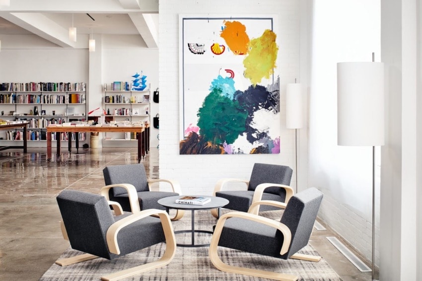 Modern office lounge with gray chairs, a round table, abstract art on white wall, shelves with books, and bright lighting.