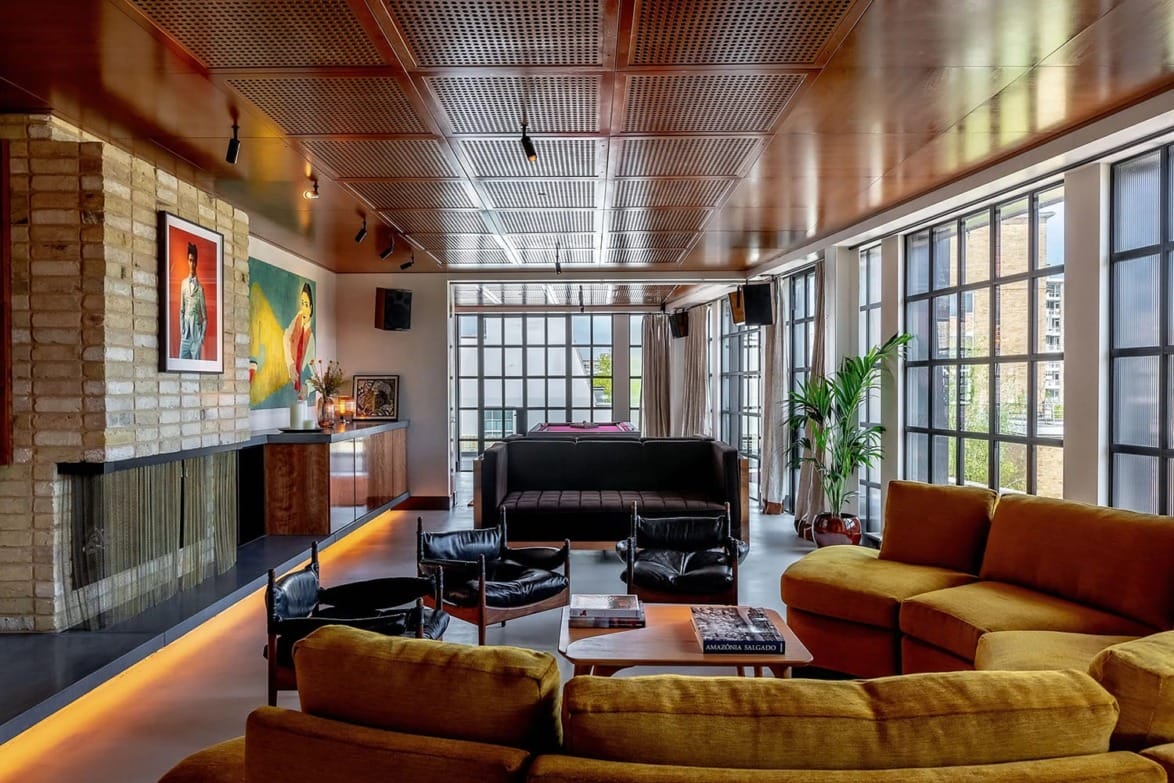 Modern living room with large windows, mustard and black sofas, artworks on walls, and a billiard table in the background.