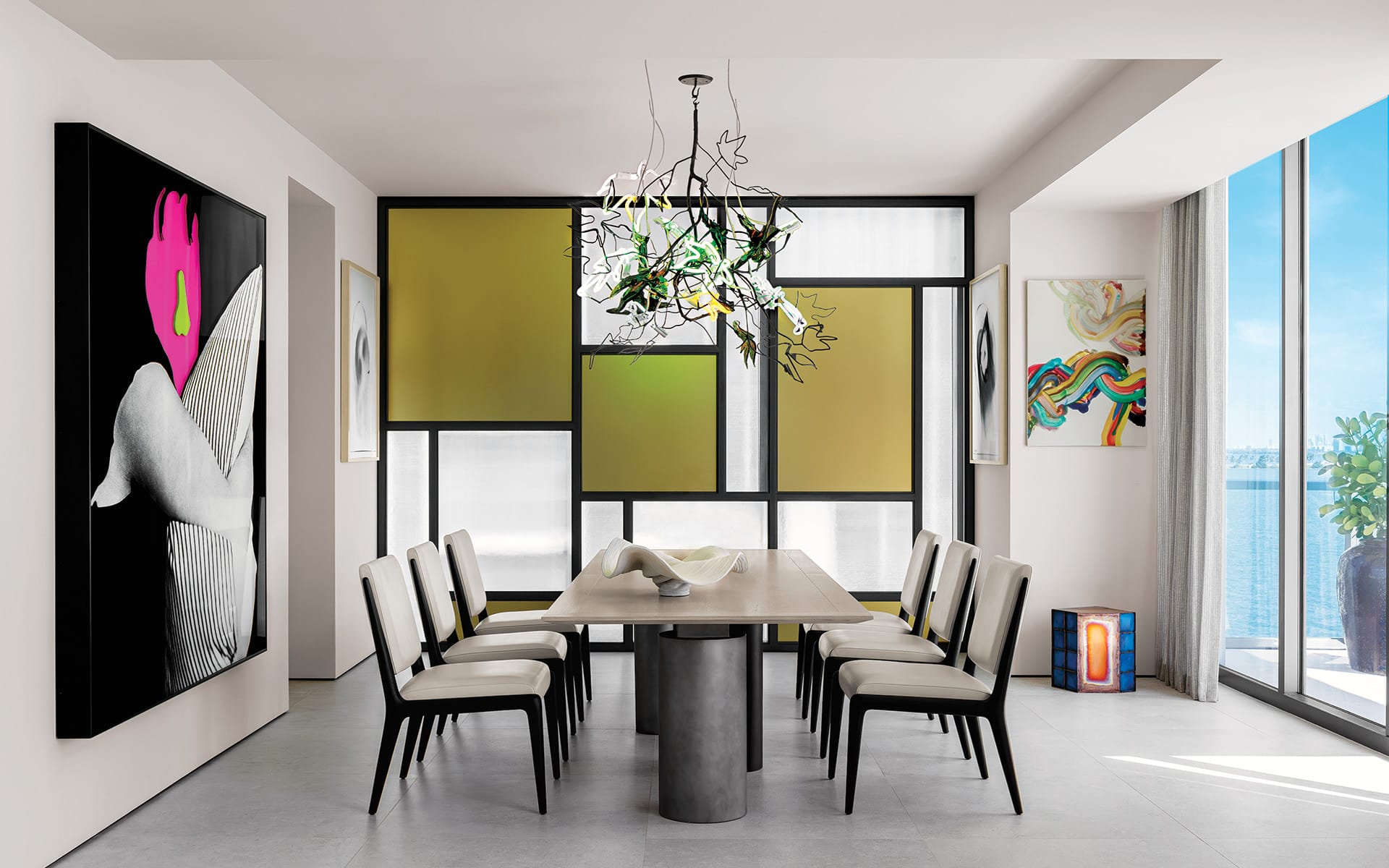 Modern dining room with artistic decor, featuring a long table, unique chandelier, and large window with a scenic view.