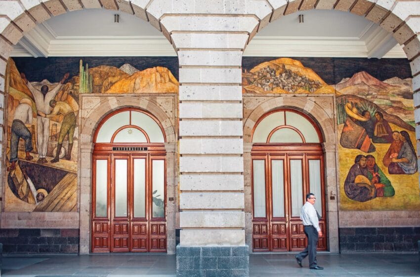 Man walking past large, colorful murals on a building with arched doorways and stone columns in an outdoor corridor.