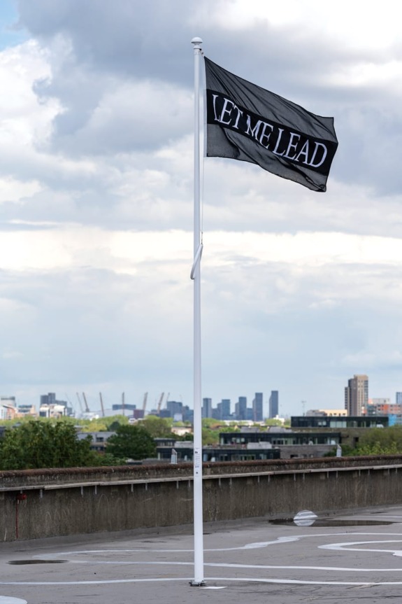 Black flag reading "LET ME LEAD" waving on a rooftop with a city skyline and cloudy sky in the background.