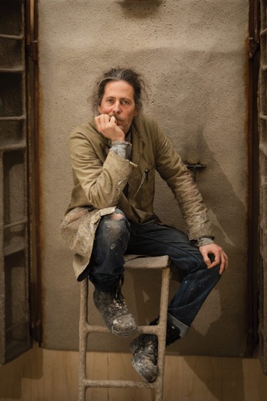 Person sitting on a ladder wearing a paint-splattered outfit, surrounded by textured walls.