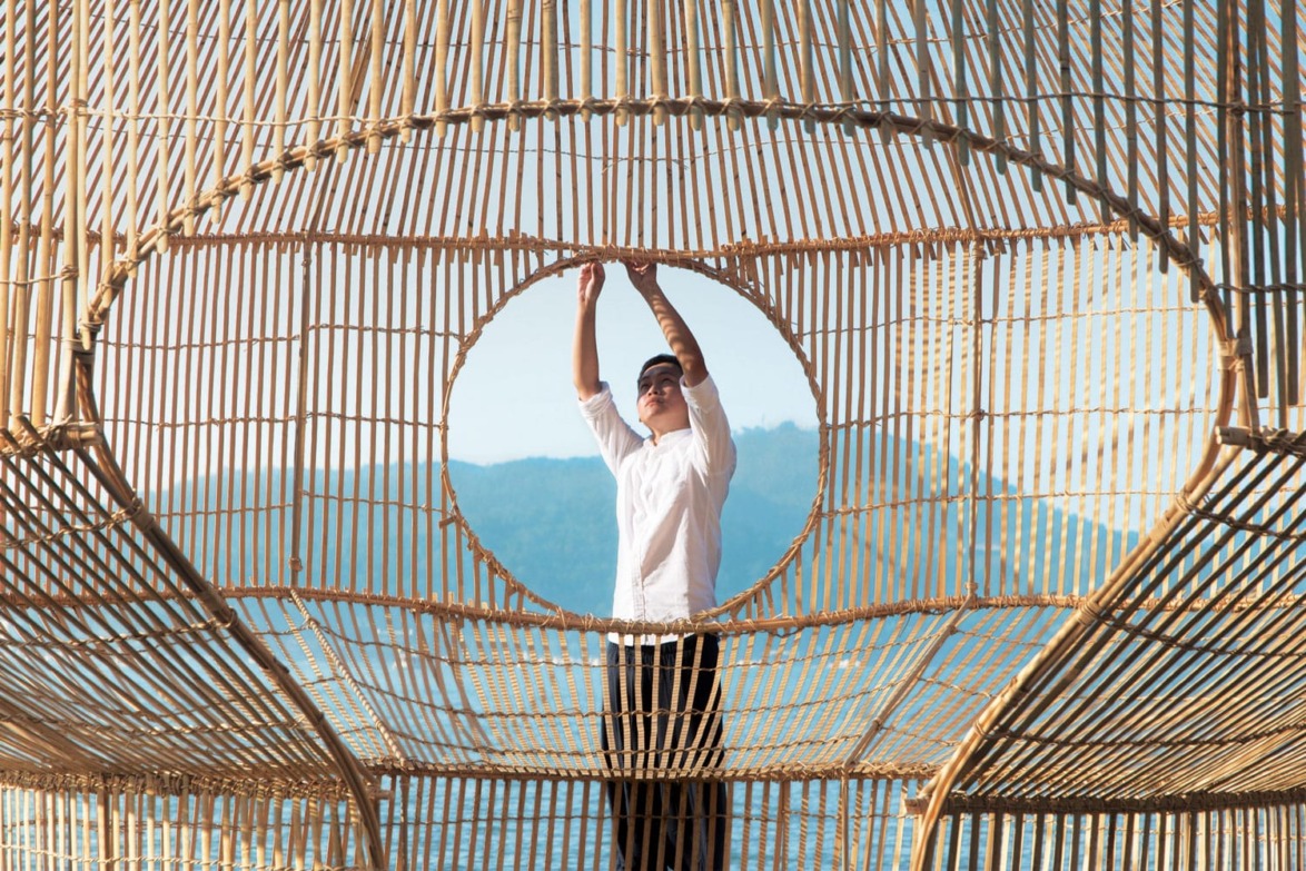 Person standing in a unique bamboo structure, adjusting the design with a mountain landscape in the background.