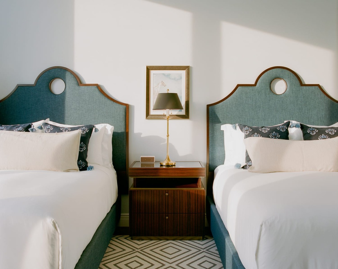 Elegant hotel room with two neatly made beds, decorative pillows, a nightstand, lamp, and framed artwork on the wall.