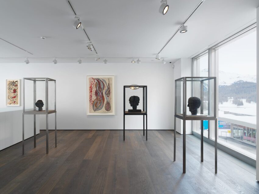 Modern art gallery with sculptures in glass cases, abstract paintings on white walls, and large windows with a snowy landscape view.