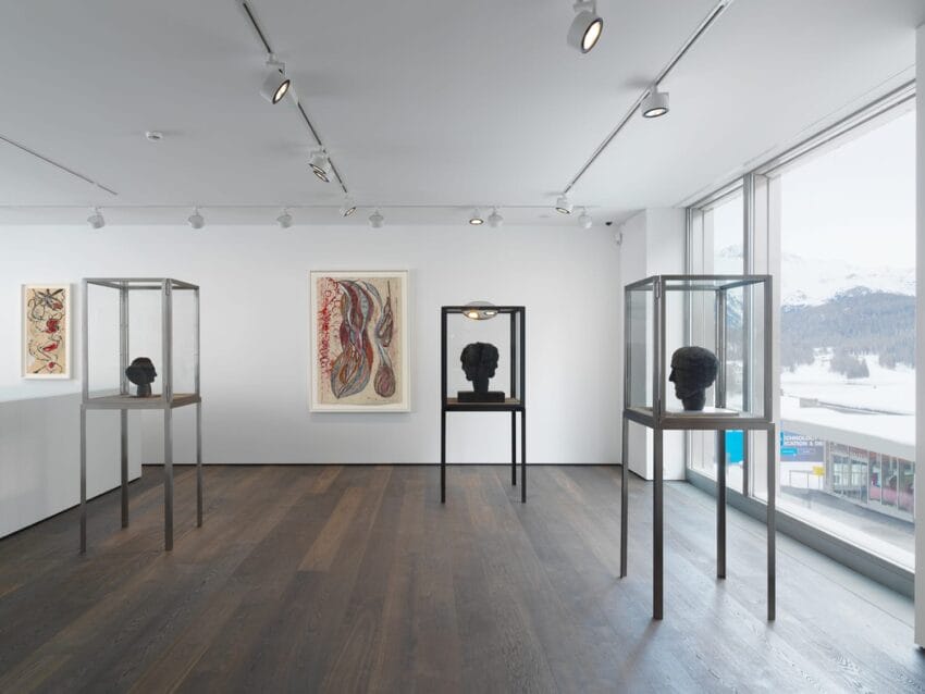 Modern art gallery with sculptures in glass cases, abstract paintings on white walls, and large windows with a snowy landscape view.